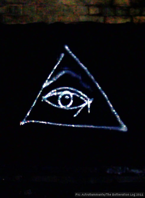 Graffito of an eye surrounded by a triangle in silver paint on a black viaduct wall.