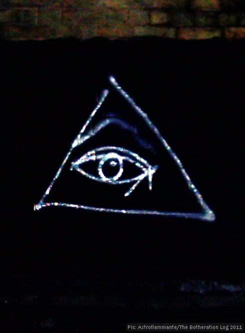 Grafitto of an eye surrounded by a triangle in silver paint on a black viaduct wall.